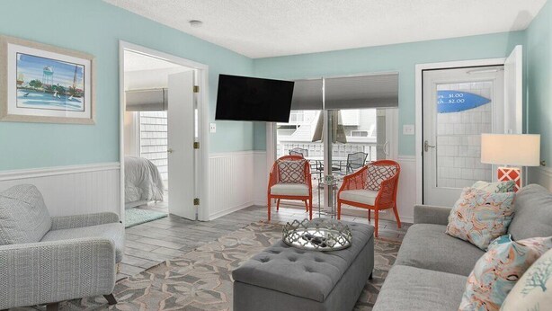 A living room with a grey sofa, a grey armchair, a grey ottoman, and a red chair with a glass table. There is a flat-screen TV mounted on the wall and a door with a sign indicating prices. - 2 BR 2 BA Unit W/Pool and Close to Northside Park (Ocean City)