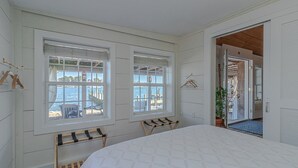 Ein Schlafzimmer mit einem großen Bett, zwei Fenstern mit Meerblick, einem Wäscheständer und einer Tür zu einem anderen Raum.