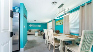 A dining area with a wooden table and chairs, a fish wall decoration, and a kitchen visible in the background. - Seaglass Inn at the Beach Shack  Just one block from the beach! (Englewood)