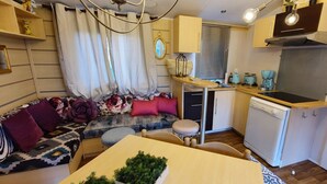 A compact living space with a sofa, a dining table, a small kitchen area including a stove and a dishwasher, and a window with blinds. - Independent cabins Las Rosas (Pollos Valladolid)