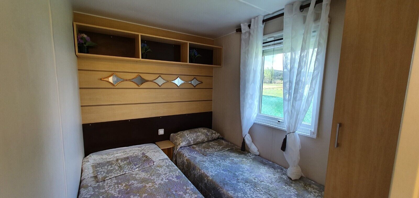 Een compacte slaapkamer met twee bedden, een houten hoofdbord met decoratieve spiegels en een raam met dunne gordijnen.