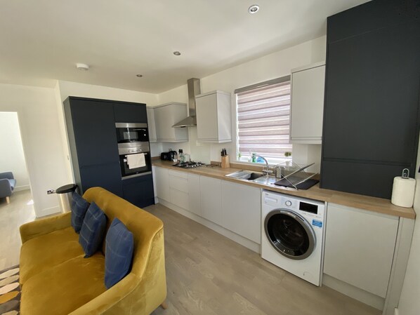 A modern kitchen with a built-in oven, microwave, and washing machine. There is a yellow sofa with blue cushions in the living area.