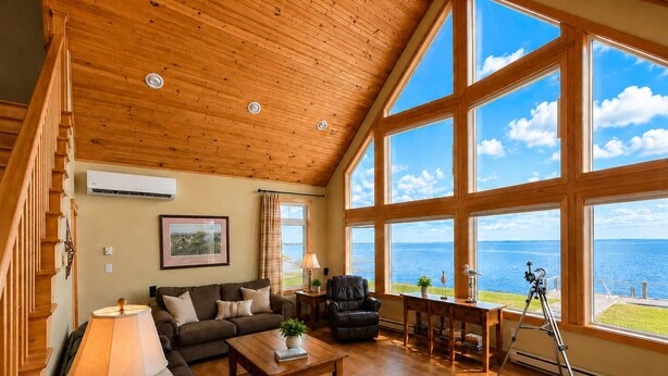 Un salon spacieux avec un plafond en bois, de grandes fenêtres offrant une vue sur la mer, un canapé, une table basse et un trépied.