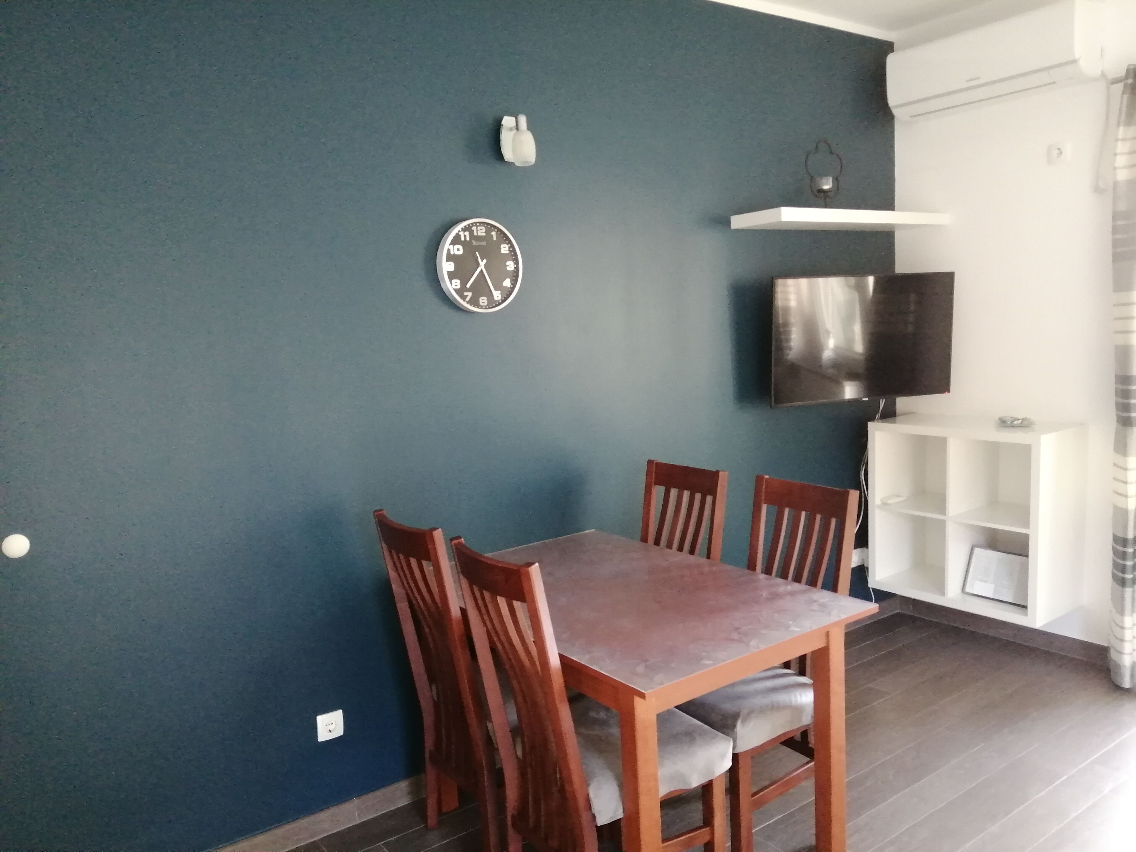 Un coin repas avec une table et des chaises en bois, une horloge au mur, un téléviseur fixé au-dessus d’une étagère blanche et un mur bleu.