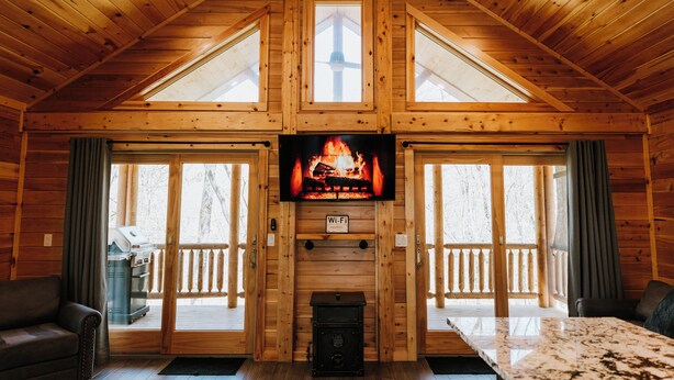 A log cabin interior with a fireplace, a television mounted above it, and a sliding glass door leading to a deck. - Cozy cabin for up to 6 guests - Hocking Hills - Firepit -Hot Tub - Wifi (Logan)