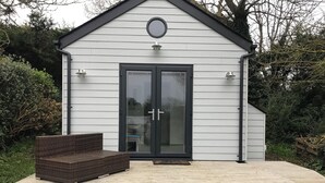 Un petit bâtiment blanc, avec une porte sombre et une fenêtre ronde au-dessus. Un banc en osier se trouve devant la porte.