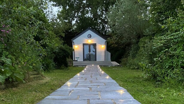 Un chemin de pierres mène à un petit bâtiment doté d’une horloge et de luminaires, le tout étant entouré de verdure.