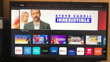 A hotel TV screen displaying a streaming service interface with various channels and a prominent advertisement for "Steve Carell: Irresistible."
