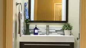 A bathroom with a dark wood vanity, a white countertop, a mirror, a towel rack, a green plant, and a blue soap dispenser.