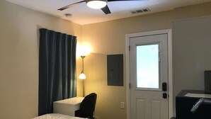 A hotel room with a bed, a desk, a chair, a desk lamp, a ceiling fan, a door, and a minibar.