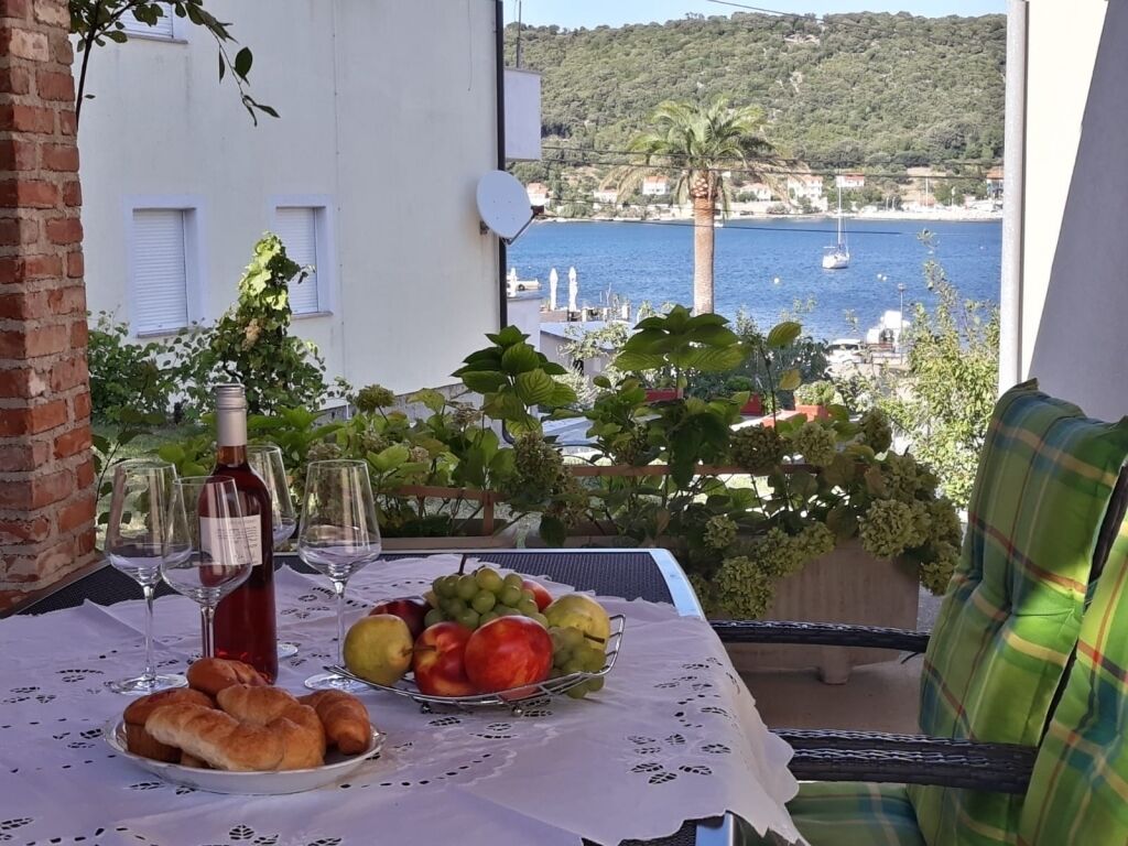 Un tavolo da balcone con una bottiglia, bicchieri, frutta e dolci, con vista sulla costa, edifici e imbarcazioni.