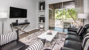 Una sala de estar moderna con un televisor de pantalla plana, una mesa de centro blanca, un sofá de cuero negro y un cojín a rayas.