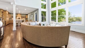 A spacious living area with a large curved sofa, a kitchen with wooden cabinets, and large windows offering a view of greenery and a body of water. - Private Lakefront Escape, Launch boat from dock, swim island, hot tub, game room (Berrien Springs)