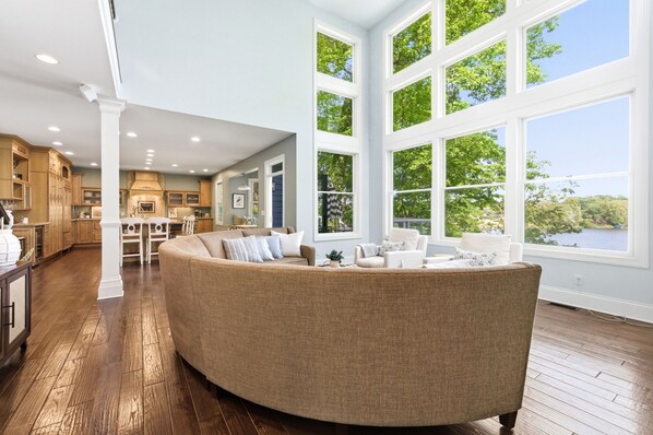 A spacious living area with a large curved sofa, a kitchen with wooden cabinets, and large windows offering a view of greenery and a body of water. - Private Lakefront Escape, Launch boat from dock, swim island, hot tub, game room (Berrien Springs)