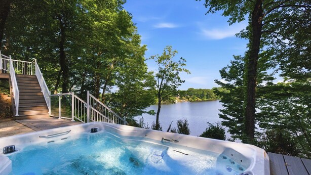 A hot tub surrounded by trees and a lake, with a wooden deck and stairs leading up to it. - Private Lakefront Escape, Launch boat from dock, swim island, hot tub, game room (Berrien Springs)