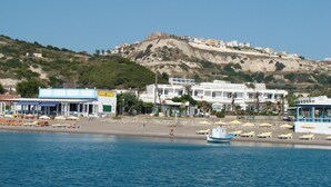 Un pueblo costero con una colina destacada, cafés frente a la playa y un barco amarrado cerca de la orilla.
