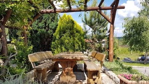 Een houten eettafel buiten met banken, omringd door weelderige begroeiing en een pergola.