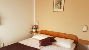Een net opgemaakt bed met wit en bruin beddengoed, een klein nachtkastje met een vaas bloemen en een ingelijst schilderij aan de muur.