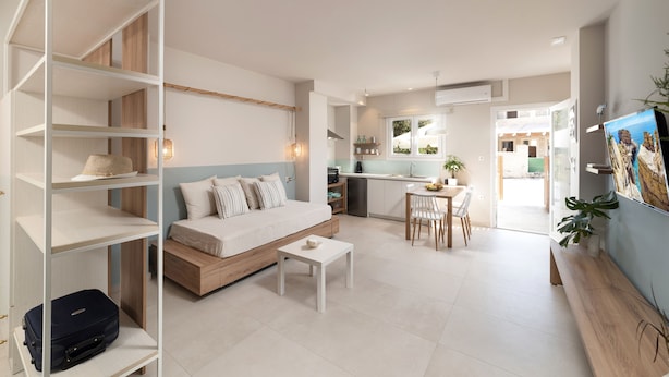 Un salon moderne et bien éclairé, avec un canapé, une petite table et une bibliothèque. On aperçoit à l’arrière-plan un coin cuisine avec une table à manger et des chaises.
