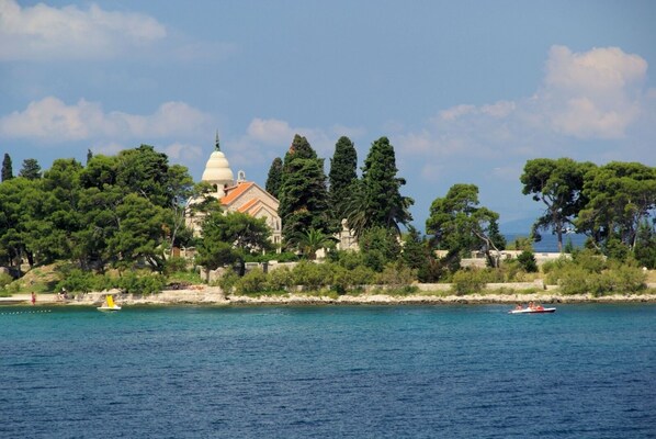 Vista costera con un edificio destacado de cúpula blanca, posiblemente una iglesia o lugar histórico, rodeado de abundante vegetación y un mar tranquilo.