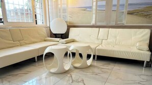 Un salon moderne avec un canapé d’angle beige, deux petites tables rondes et une grande fenêtre donnant sur la plage.