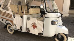 Un véhicule ancien à trois roues, de couleur blanche, doté d’une capote, de sièges en cuir beige et d’un emblème héraldique.