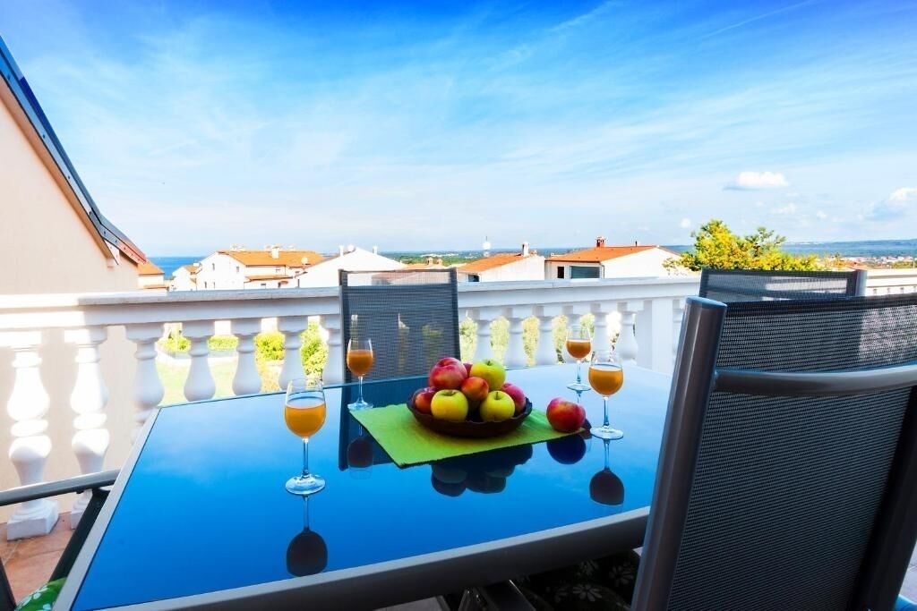 Une table de balcon avec un panier de fruits et des verres de jus d’orange, donnant sur un quartier résidentiel.