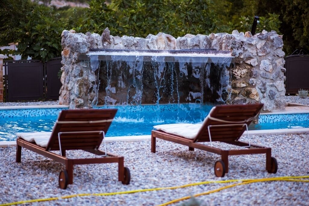 Uma área de piscina com duas espreguiçadeiras de madeira, uma cascata de pedra e uma piscina azul cristalina rodeada por pedrinhas.