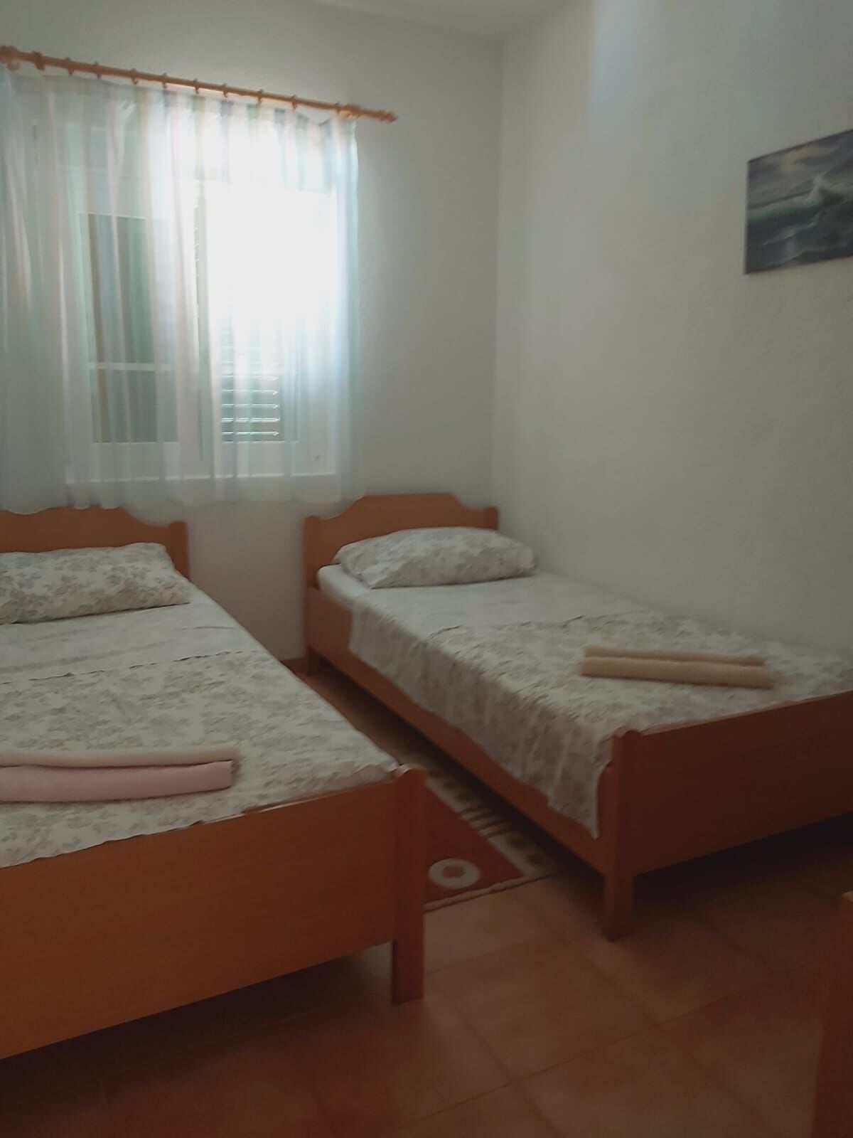 A bedroom with two single beds, a window with sheer curtains, and a painting on the wall.