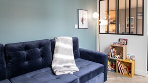 Salón con un sofá azul oscuro, una manta blanca, un cuadro en la pared y una estantería con libros y juguetes infantiles.