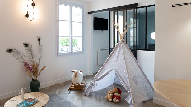 Una habitación acogedora con un ventanal grande, piso de madera, una mesita con un libro, un jarrón decorativo, un tipi y peluches.
