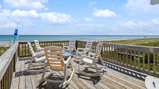 Eine Holzterrasse mit weißen Schaukelstühlen und einem Tisch, mit Blick auf einen Strand und einen klaren Himmel.