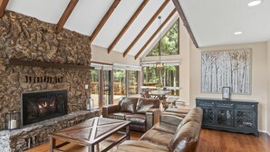 A spacious living room with a stone fireplace, leather furniture, and a large window offering a view of the outdoors. - Peaceful Forest Retreat· Hot Tub · Sauna · Views (Flagstaff)