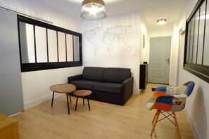 A modern living room with a black sofa, two small wooden tables, a colorful chair, a world map on the wall, and a window with blinds. - Villa Jean Julien - Les Cascades - Appartement T3 - 2 Chambres - 6 Pers (Mont-Dore)