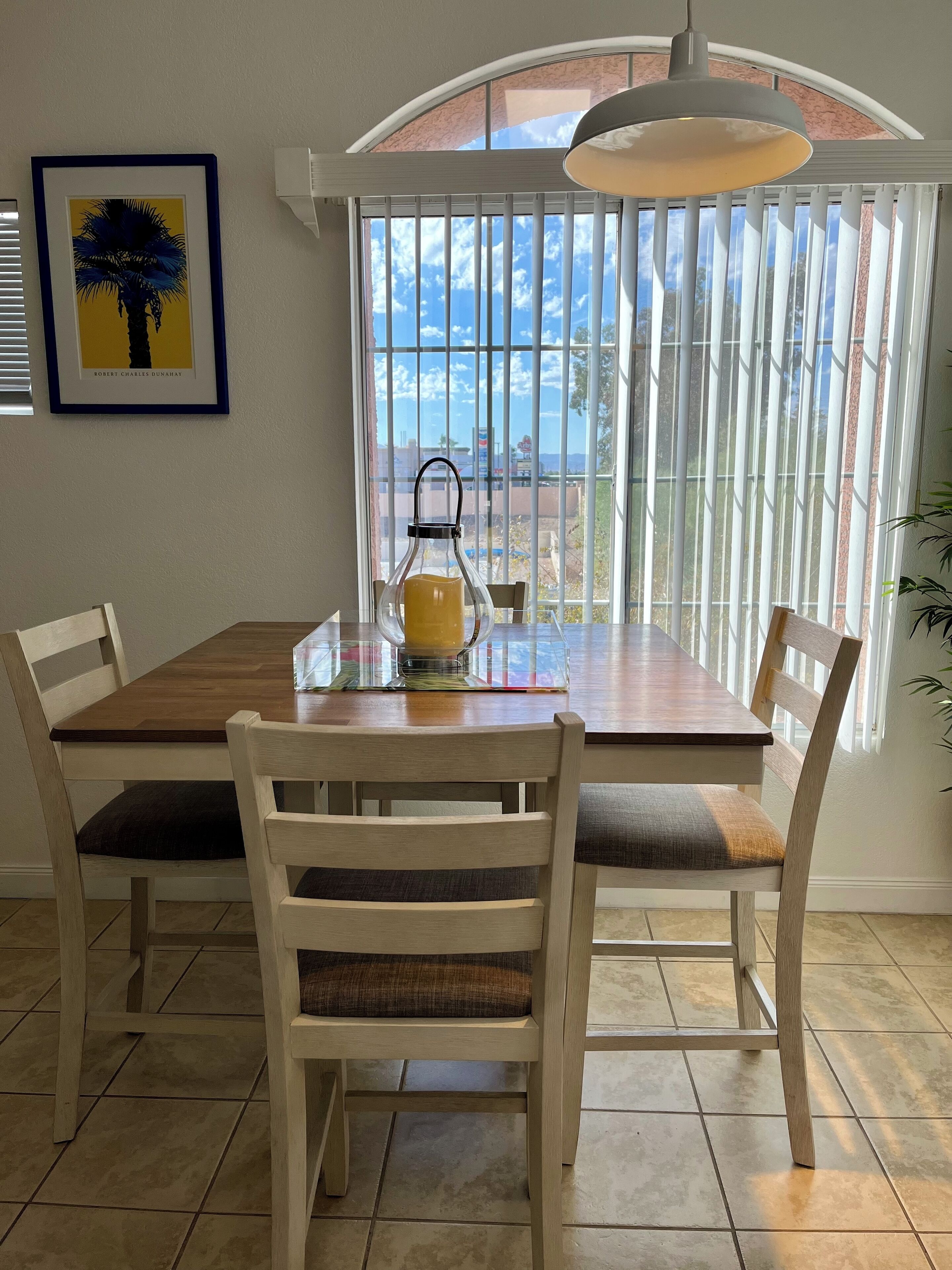 A dining area with a wooden table and chairs, a clear view of the sky through large windows, and a framed artwork on the wall.