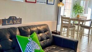 Ein Wohnzimmer mit einer schwarzen Ledersofa, zwei Kissen, einem Schild mit der Aufschrift 'Zum See', gerahmten Bildern an der Wand, einem Essbereich mit Tisch und Stühlen sowie einer Lampe.