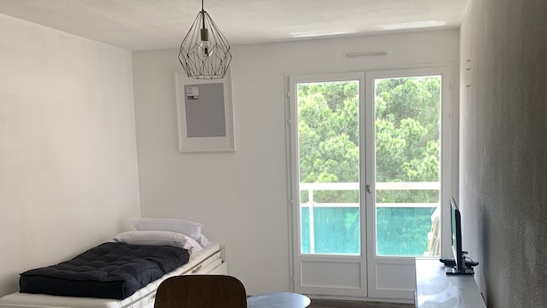 Een kleine kamer met een bed, een houten tafel, een stoel, een klein rond tafeltje, een bureau, een plafondlamp en een deur naar een balkon met uitzicht op groen.