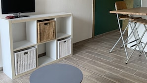 Een moderne woonkamer met een flatscreen televisie op een wit schap, rieten manden, een ronde tafel met een geweven vloerkleed, en een eethoek met een metalen stoel en tafel.