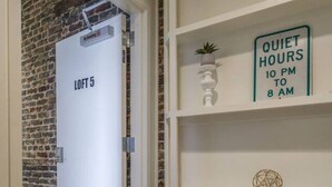 Un pasillo con pared de ladrillo, una puerta blanca con la leyenda 'LOFT 5', un cartel de 'HORAS DE SILENCIO' y una estantería con una planta pequeña y un adorno.