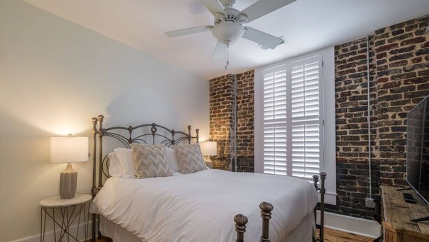 Un dormitorio con pared de ladrillo, cama con estructura metálica, ventilador de techo y ventana con contraventanas.