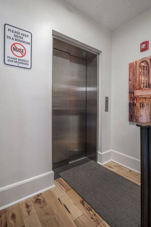 Un ascenseur moderne aux finitions métalliques, un tapis gris à l’entrée et une pancarte fixée au mur demandant aux usagers de faire le moins de bruit possible.