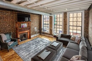 Un salon avec un mur en briques, une cheminée, un canapé et de grandes fenêtres.