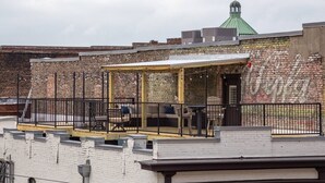 Una terraza en la azotea con zona de estar cubierta, un muro de ladrillo con grafitis y un edificio con techo verde al fondo.
