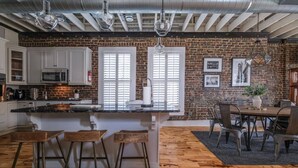 Una cocina con pared de ladrillo, armarios blancos, una isla de madera con taburetes y un comedor con mesa y sillas.