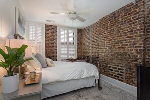 Une chambre avec un mur en briques, un lit avec une literie blanche, un ventilateur de plafond et une fenêtre avec des stores.