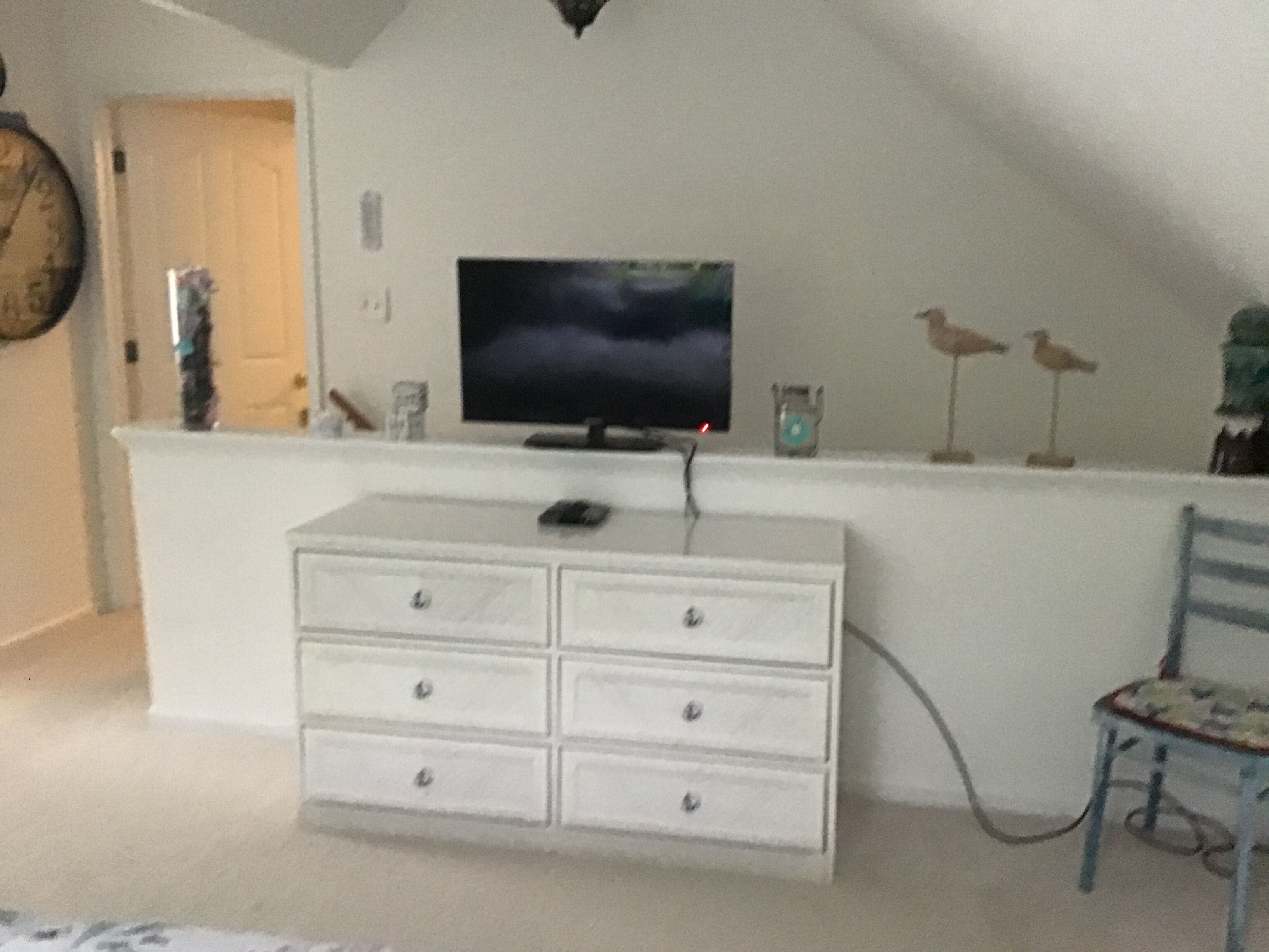 Een kamer met een witte dressoir, een televisie op een standaard, een klok aan de muur, en een stoel met een gestreept zitvlak.