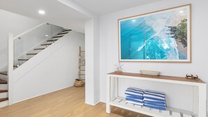 Un intérieur moderne avec un escalier en bois, un tableau représentant une vue sur l’océan, encadré, et une console sur laquelle sont posées des serviettes.