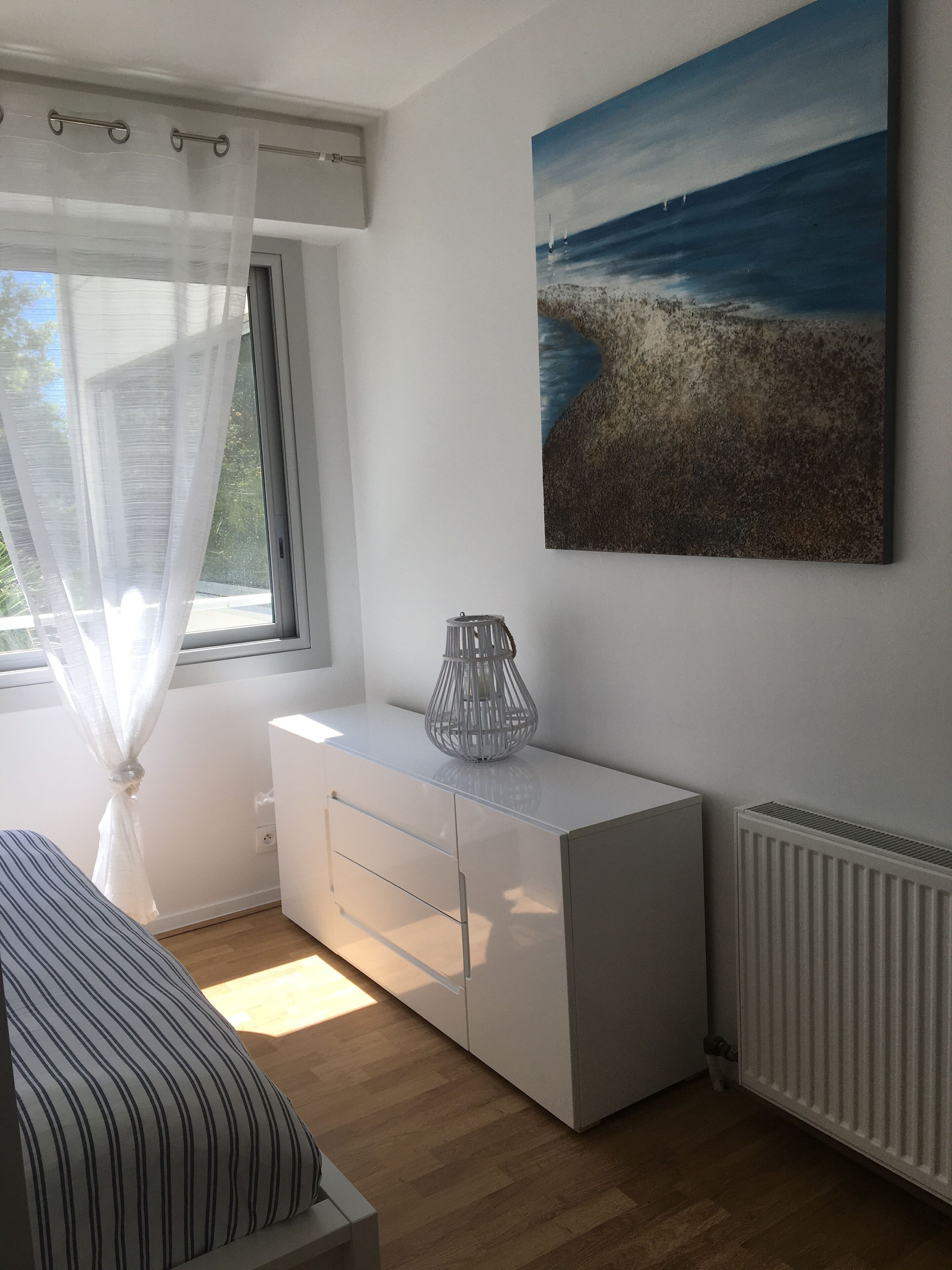 A bedroom with a bed, a bedside table, a window with sheer curtains, a white dresser, a radiator, and a painting of a beach scene.