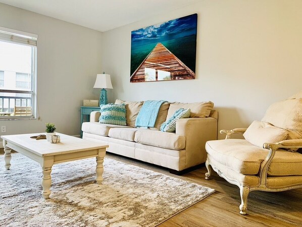 A living room with a beige sofa, a white coffee table, a blue lamp, and a painting of a pier. - Condo Close To The Beach (Venice)