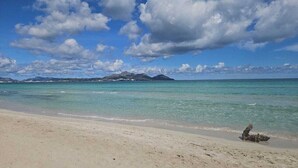 Una platja amb aigua turquesa transparent, sorra daurada i muntanyes al capvespre sota un cel amb núvols dispersos.